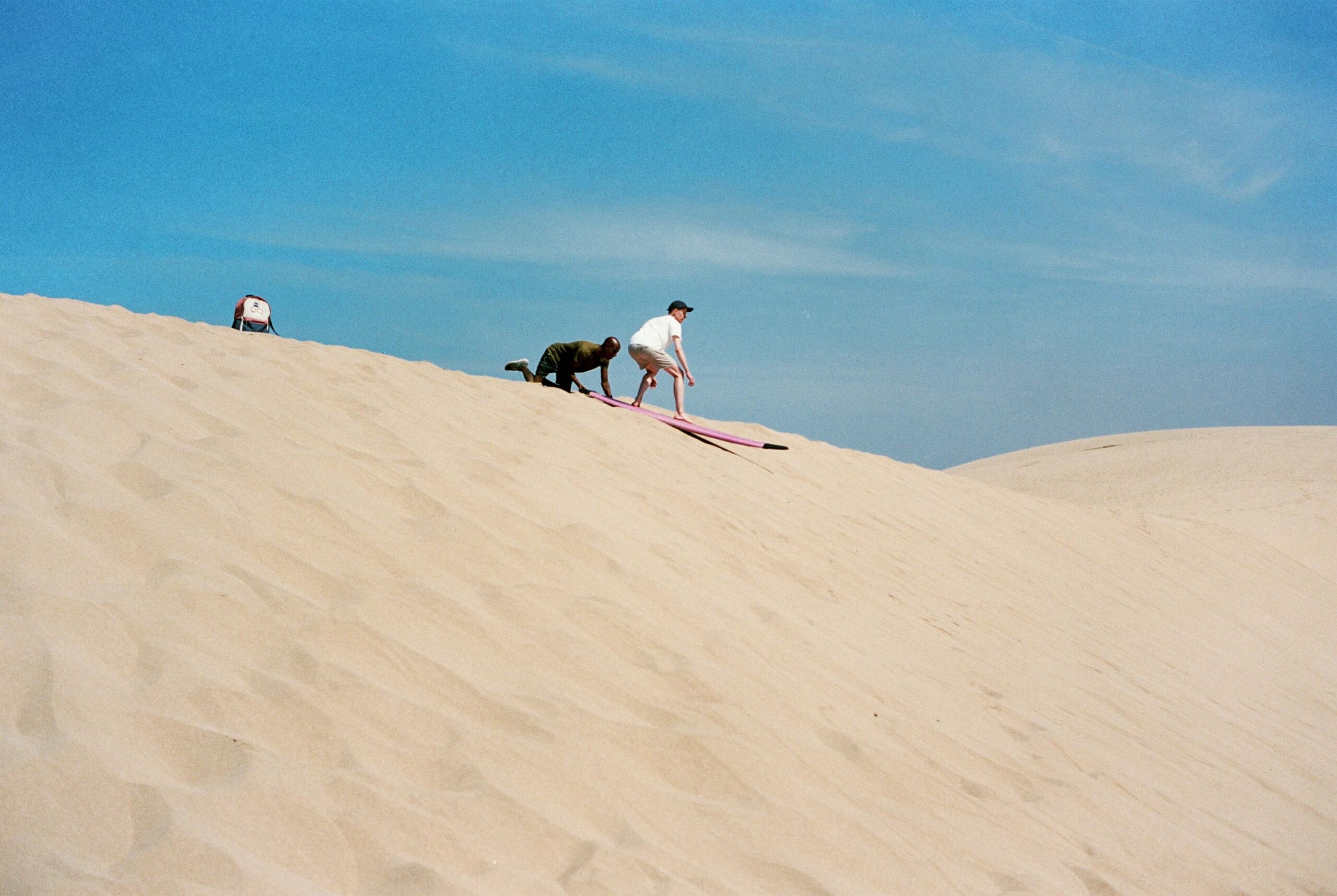 Two people enjoying a sandboarding adventure on the dunes of Morocco under a clear blue sky.